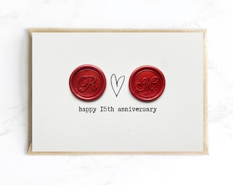 Puede incluir: Tarjeta crema en un sobre marrón. Presenta dos sellos de cera rojos con iniciales, un corazón dibujado a mano y el texto "happy 15th anniversary". El fondo es una superficie de mármol blanco. Ideal para un 15 aniversario.
