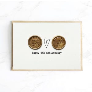 May include: A white card with a brown envelope features two gold-colored wax seals with the letters "R" and "S", a heart, and the text "happy 8th anniversary". The card is on a white surface.