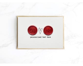 Op de afbeelding: Een witte rechthoekige kaart met twee ronde rode waszegels met reliëfletters, gescheiden door een handgetekend zwart hart. Onder de zegels staat de tekst "personalised text here" gedrukt in een zwart lettertype in typemachinestijl.