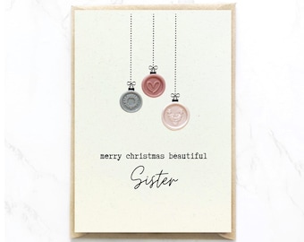 Op de afbeelding: Een crèmekleurige wenskaart met drie hangende waszegelornamenten in grijs, oudroze en lichtroze, met de tekst merry christmas beautiful Sister in zwarte typemachine- en scriptlettertypes op de voorzijde van de kaart gedrukt.