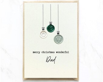 Op de afbeelding: Een crèmekleurige wenskaart met drie hangende waszegelornamenten in wit, donkergroen en lichtgroen. De zegels tonen een krans, een hart en een hertenkop. De tekst luidt merry christmas wonderful Dad in zwarte inkt.