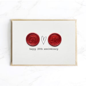 May include: A white card with a brown border features two red wax seals with initials and a hand-drawn heart. The text "happy 16th anniversary" is printed below the seals. A simple and elegant design.