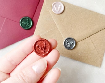 Op de afbeelding: Een rond, metallic koperkleurig waszegel met een reliëf van een hertenkop. Extra waszegels in groen, zilver en lichtroze zijn bevestigd op donkerrode en bruine enveloppen, die het hertenmotief in verschillende kleuren laten zien.