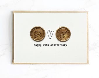Op de afbeelding: Een witte kaart met een bruine envelop toont twee gouden zegels met de letters "R" en "S", een handgetekend hart en de tekst "happy 19th anniversary" in het zwart. De kaart is een romantische jubileumgroet.