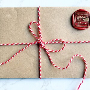 Puede incluir: Un sobre de papel marr&oacute;n atado con hilo rojo y blanco. Un sello de cera rojo con letras doradas est&aacute; adherido en la esquina superior derecha. El hilo est&aacute; atado en un lazo.