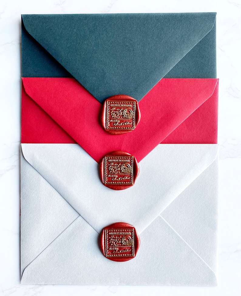 Puede incluir: Tres sobres en gris, rojo y blanco, sellados con sellos de cera rojos. Los sellos tienen un dise&ntilde;o dorado en relieve. Los sobres est&aacute;n dispuestos en un patr&oacute;n superpuesto, con el sobre gris en la parte superior.