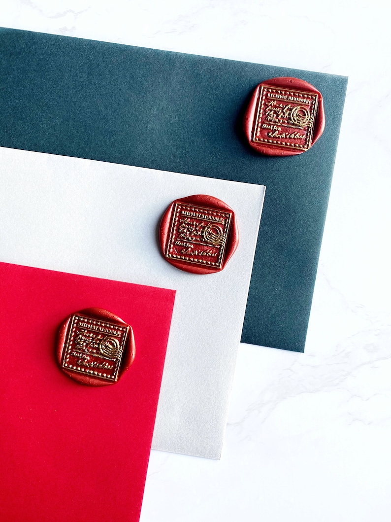 Puede incluir: Sellos de cera rojos con letras doradas est&aacute;n adheridos a sobres rojos, blancos y azul oscuro. Los sellos tienen un dise&ntilde;o rectangular con las palabras "DELIVERY CONFIRMED" y otro texto. Los sobres est&aacute;n dispuestos sobre una superficie de m&aacute;rmol blanco.
