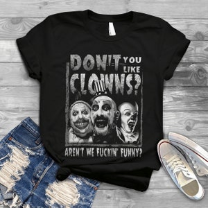 May include: Black t-shirt with a graphic of three unsettling clowns and the text "DON'T YOU LIKE CLOWNS? AREN'T WE FUCKIN' FUNNY?". The design uses grey and white tones for a vintage, distressed effect. The shirt is shown on a wooden surface.