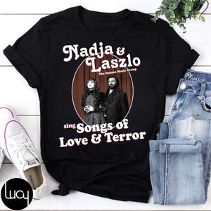 Könnte beinhalten: Schwarzes T-Shirt mit dem Text "Nadja & Laszlo" in einer Retro-Schriftart und einem Bild von zwei Personen in einem Kreis. Darunter steht "sing Songs of Love & Terror". Das Shirt wird mit Jeans und Turnschuhen kombiniert.