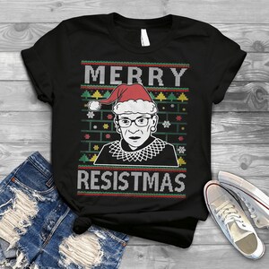 May include: Black t-shirt with a white and red Christmas sweater design featuring a portrait of Ruth Bader Ginsburg wearing a Santa hat. The text "Merry Resistmas" is printed below the portrait.