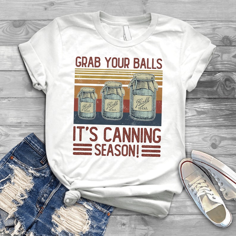 May include: White t-shirt with a vintage-style graphic that says "Grab Your Balls It's Canning Season!" with three Ball canning jars in the design.