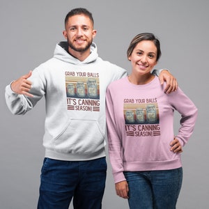 May include: Two people wearing hoodies with a vintage-style graphic that says "Grab your balls it's canning season!" The graphic features three jars of preserved food. The person on the left is wearing a white hoodie and the person on the right is wearing a pink hoodie.