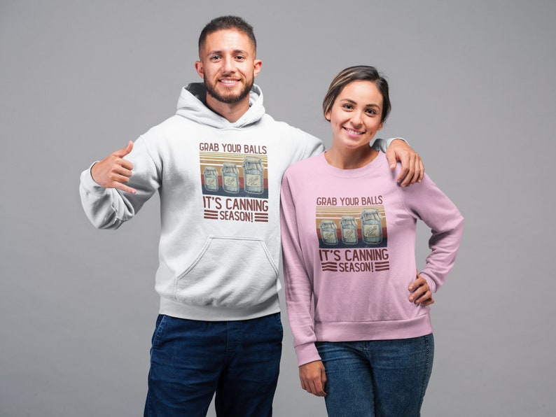 May include: Two people wearing hoodies with a vintage-style graphic that says "Grab your balls it's canning season!" The graphic features three jars of preserved food. The person on the left is wearing a white hoodie and the person on the right is wearing a pink hoodie.