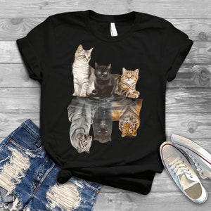 May include: Black t-shirt with a graphic of four cats reflected in water as tigers. The cats are a tabby, a black cat, a white cat, and a calico cat.