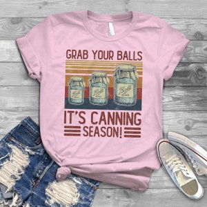 May include: Pink t-shirt with a vintage-style graphic that says "Grab Your Balls It's Canning Season!" with three jars labeled "Ball Ideal" in a retro design.