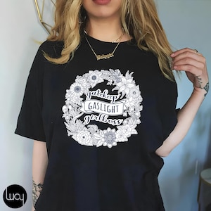 Peut inclure: T-shirt noir avec un motif de couronne de fleurs et le texte "gatekeep GASLIGHT girlboss".