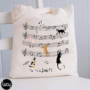 May include: Cream-colored tote bag featuring a musical staff design with playful cat illustrations. The cats, in various colors, are interacting with the musical notes. The bag has a simple design with a handle for carrying.