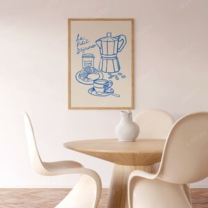 May include: A blue line drawing of a coffee pot, a cup of coffee, a croissant, and a jar of jam on a cream background. The text "Le Petit D&eacute;jeuner" is written in blue above the drawing. The drawing is framed in a light wood frame.