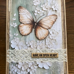 Puede incluir: Un diario decorativo con un diseño de mariposa y flores. La portada presenta una mariposa grande y detallada con alas marrones y beige, rodeada de flores blancas. También se incluye una tira de encaje y un letrero de madera que dice "HAVE AN OPEN HEART".