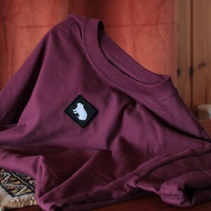 May include: A maroon t-shirt with a white bear silhouette patch on the front.