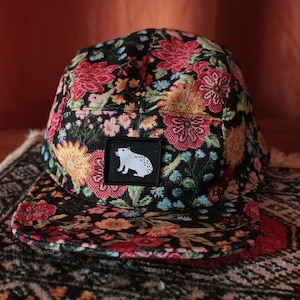 Peut inclure: Une casquette noire à 5 panneaux avec un motif floral dans les tons rouge, jaune, bleu et vert. La casquette a un patch blanc avec une silhouette noire d'un ours sur le devant.
