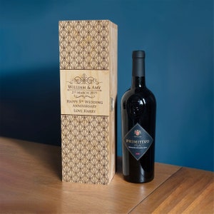 Puede incluir: Una caja de madera para vino con un patrón geométrico y un mensaje personalizado. El mensaje dice "William & Amy 2nd March 2019 Happy 5th Wedding Anniversary Love Harry". Una botella de vino tinto está al lado de la caja.