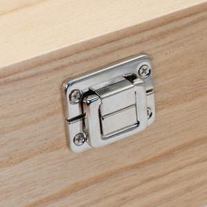 May include: A close-up of a silver metal latch on a wooden box. The latch has a rectangular shape with a small, rectangular opening in the center. The latch is attached to the box with two screws.