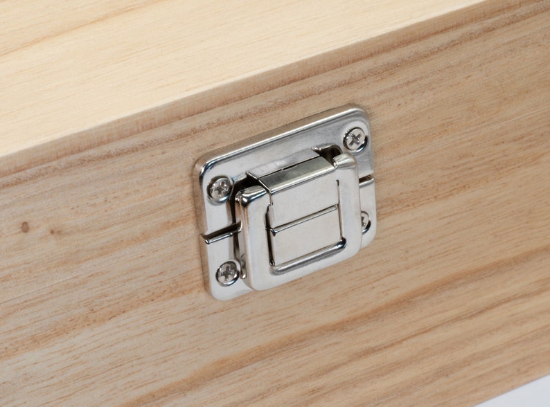 May include: A close-up of a silver metal latch on a wooden box. The latch has a rectangular shape with a small, rectangular opening in the center. The latch is attached to the box with two screws.