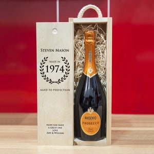 Puede incluir: Una caja de regalo de madera con una botella de prosecco en su interior. La caja está grabada con el texto "Steven Mason Made in 1974 Aged to Perfection" y "Hope you have a great one Love Amy & William".