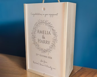 May include: A wooden box with a rope handle, personalized with the names Amelia & Harry, the date 22nd October 2024, and the message "Congratulations on your engagement." The box is decorated with a wreath of leaves and the text "Love from Rachael, Simon & Family xxx."