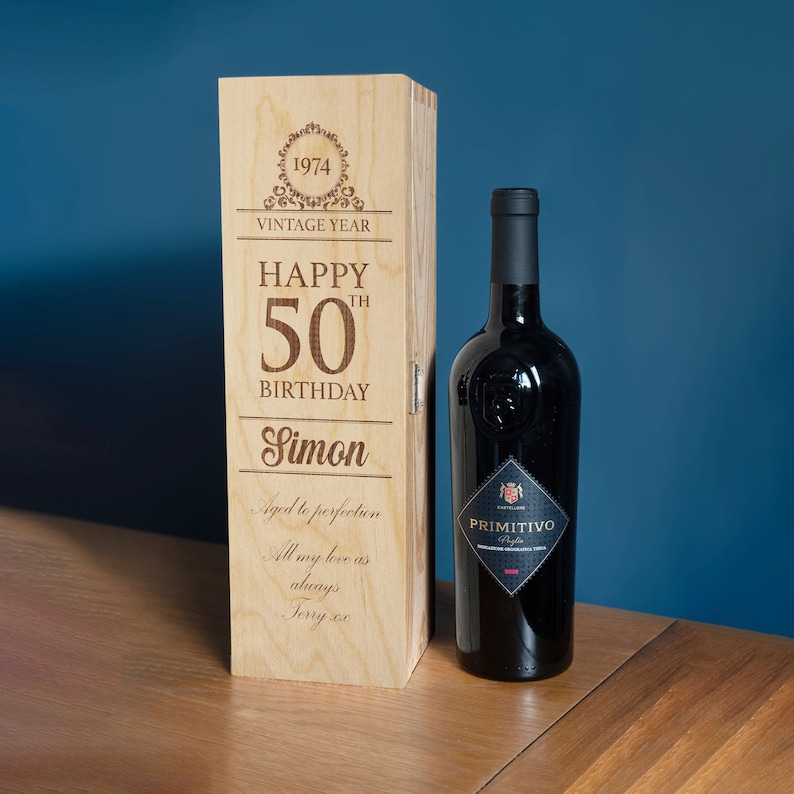 May include: A wooden wine box with a personalized message for a 50th birthday. The box is engraved with the text "Happy 50th Birthday Simon" and "Aged to perfection". The box is closed and a bottle of wine is visible in the background.