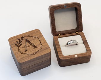 May include: Two wooden ring boxes, one closed and one open. The closed box has a geometric design with the letter "A" and the name "Amelia" engraved on the lid. The open box displays a silver ring with a small gem, nestled in a cream-colored interior.