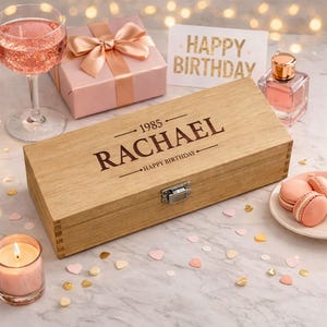 May include: A wooden wine box engraved with "RACHAEL" and "HAPPY BIRTHDAY" is displayed with a gift box, a glass of pink drink, perfume, and macarons. The box has a metal clasp. A lit candle and confetti add to the celebratory atmosphere. The scene is set on a marble surface.