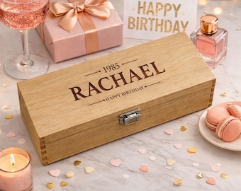 May include: A wooden wine box engraved with "RACHAEL" and "HAPPY BIRTHDAY" sits on a marble surface, surrounded by a gift box, a glass of pink drink, a perfume bottle, and macarons. The box is secured with a metal clasp. A lit candle and confetti complete the festive scene.