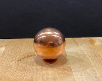 May include: A shiny copper sphere, reflecting the surrounding environment. The sphere is sitting on a light brown wooden surface.