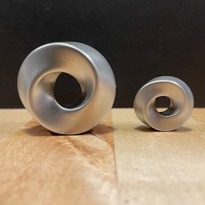 Pode incluir: Duas esculturas de metal prateado em forma de fita de Möbius, uma maior que a outra, sobre uma superfície de madeira.