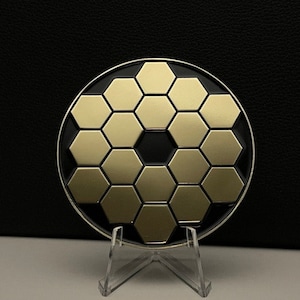 May include: A gold and black circular coin with a honeycomb pattern. The coin is on a clear plastic stand.