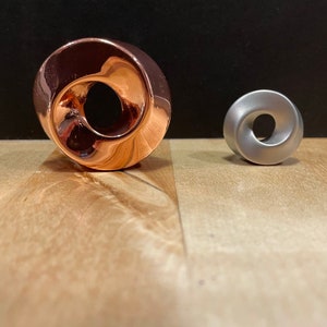 Pode incluir: Dois objetos decorativos, um é um anel de cor cobre com um furo no centro, o outro é um anel de cor prata com um furo no centro.