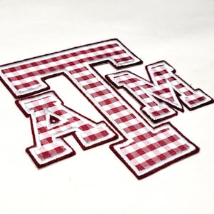 May include: An embroidered patch featuring the letters ATM in a block font. The letters are filled with a red and white gingham check pattern and are outlined with a thick, dark red border and a thin white inner border on a plain background.