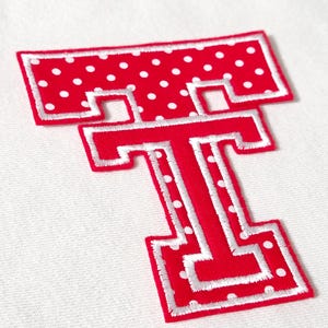 May include: A red and white embroidered letter T patch featuring a polka dot pattern on the top bar and a solid red center with a white stitched border. The letter has a block-style font design with a distinct white outline surrounding the red fabric.