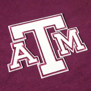 May include: A maroon fabric background featuring a large, white embroidered patch with the letters ATM in a block font. The letters are outlined with a thin maroon border, creating a distinct contrast against the white fill and the maroon textile.