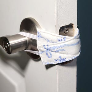 May include: A white fabric door silencer featuring a blue floral pattern with elastic bands attached to a brushed nickel door handle and latch plate. The fabric strap wraps around the handle and the door edge to prevent the door from latching shut.