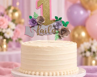 Könnte beinhalten: Ein Tortenstecker mit einer großen, gold glitzernden Eins mit rosa Rand, platziert über einem grauen Banner mit dem Namen Laila in goldener Glitzerschrift. Das Design ist mit Papierblumen in Lila- und Beigetönen sowie grünen Blättern verziert.