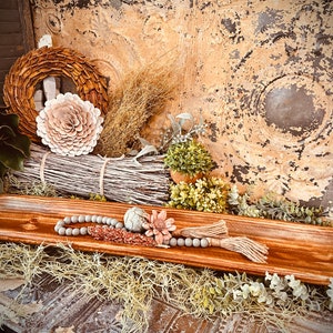 May include: A rustic wooden dough bowl filled with decorative elements. The bowl is surrounded by dried grasses, faux greenery, and a beaded garland with tassels. A wreath and a large flower are in the background, creating a farmhouse aesthetic.