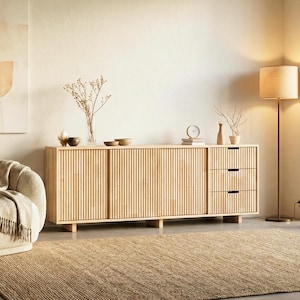 Peut inclure: Buffet en bois clair avec des détails verticaux côtelés sur les portes et les tiroirs. L'armoire a trois tiroirs sur le côté droit. Un tapis beige et un lampadaire sont dans la pièce.