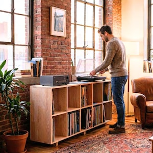 Puede incluir: Un mueble de almacenamiento de discos de madera clara con múltiples compartimentos, llenos de discos de vinilo. Un tocadiscos negro se encuentra encima, con una persona operándolo. La habitación tiene paredes de ladrillo visto y grandes ventanas.
