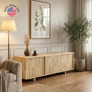 May include: A light wood media console with vertical ribbed detailing and sliding doors. A framed botanical print hangs above the console. A floor lamp with a beige shade and a potted olive tree are also in the room.