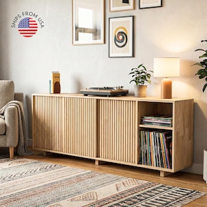 May include: A light wood-tone media console with vertical slat detailing on the sliding doors. The console features a built-in shelf for vinyl records, a turntable, and a lamp. The image includes the text "SHIPS FROM USA".