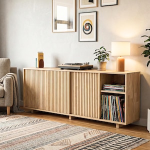 May include: A light wood-tone media console with a slatted front and sliding doors. A record player, lamp, and framed art pieces sit atop the console. A patterned rug is in the foreground. The console has a shelf for vinyl records.