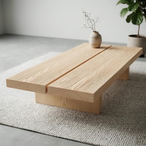 Op de afbeelding: Een lichtgekleurde houten salontafel met een rechthoekig blad dat in twee delen is gesplitst. De tafel heeft een eenvoudig, modern ontwerp met een stevige basis en staat op een grijs tapijt. Een kleine vaas met takken staat erop.